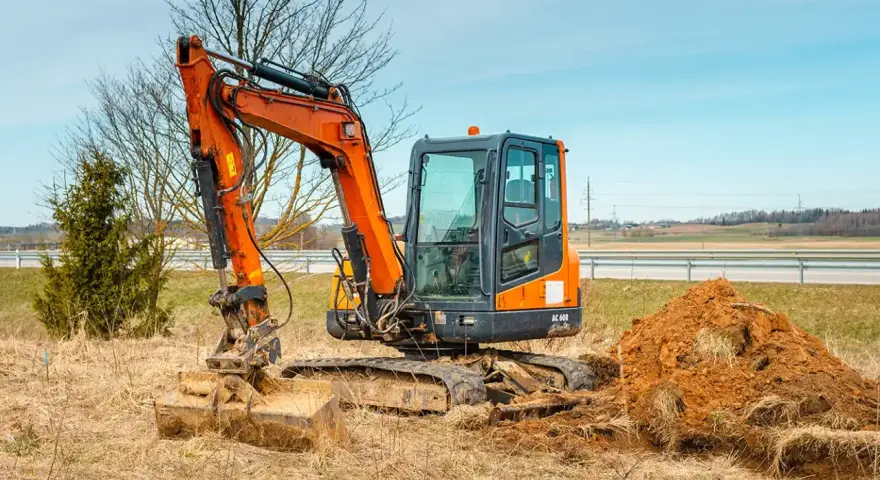 Pomarańczowa minikoparka na gąsienicach stoi na zaoranym polu, obok usypana kopa ziemi. Czas na wymianę oleju hydraulicznego w minikoparce.