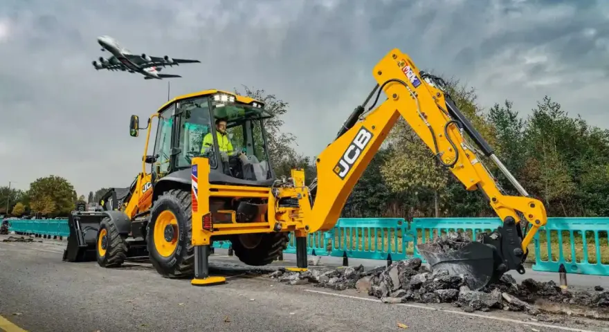Żółta koparka JCB pracuje na drodze, a nad nią przelatuje samolot. Ciekawe, ile pali koparka, gdy pracuje tak wydajnie.