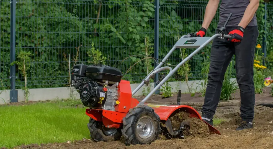 Osoba w rękawiczkach obsługuje czerwoną glebogryzarkę spalinową, spulchniając ziemię w ogrodzie. Dowiedz się, jak używać glebogryzarki spalinowej.