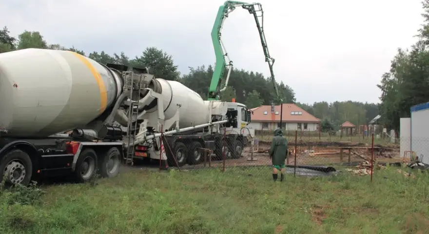 Betoniarka z wysięgnikiem pompuje beton. Pracownik w zielonej kurtce obserwuje. To ważny etap budowy, a czyszczenie betoniarki po pracy, jak usunąć zaschnięty beton z betoniarki, jest kluczowe.