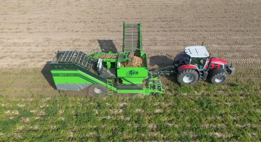 Kombajn AVR Spirit 9200 zbiera ziemniaki na polu. Czerwony traktor ciągnie maszynę, która wypełnia się plonami.