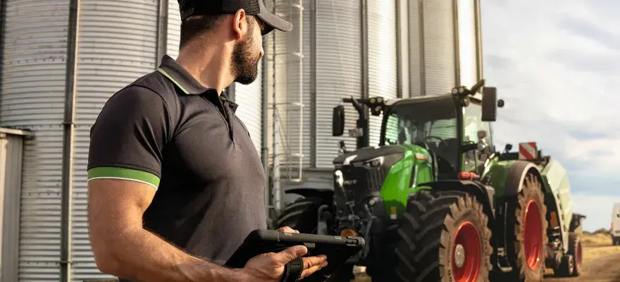 Rolnik z tabletem przy silosach, obok zielony traktor Fendt. W tle biała furgonetka.