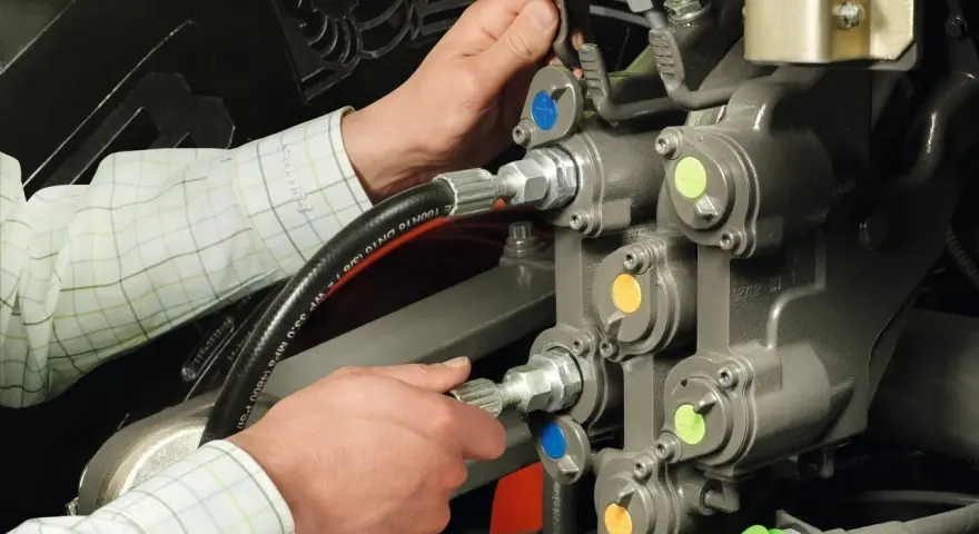 Ręce mechanika pracują przy skomplikowanym układzie hydraulicznym. Widać węże i zawory z kolorowymi oznaczeniami.