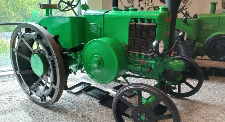 Zielony, zabytkowy Ursus, najstarszy model, z wielkimi kołami i kierownicą, stoi w muzeum.