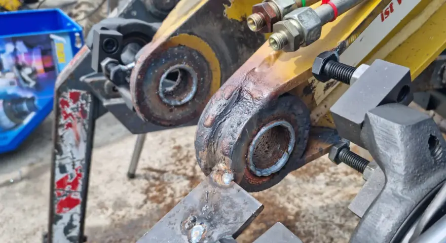 Naprawa tulejowania koparki. Widoczne elementy hydrauliczne i zardzewiałe sworznie.