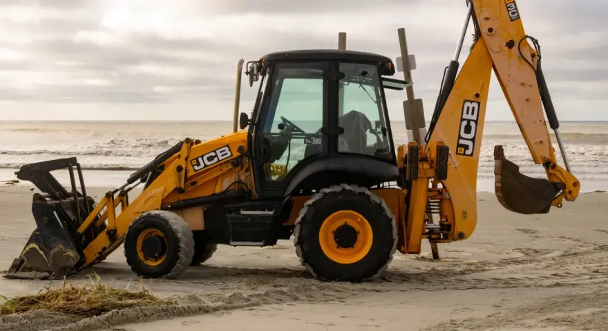 Żółta koparko-ładowarka firmy JCB stoi na piaszczystej plaży, z widokiem na morze i zachmurzone niebo.