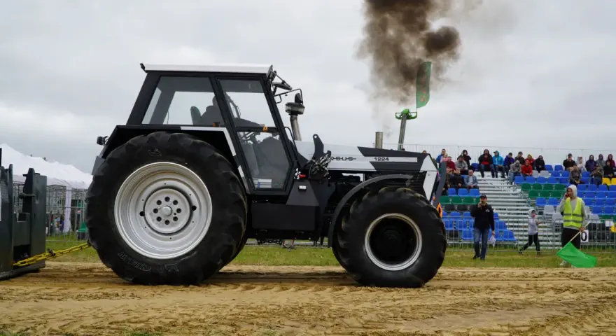Ciężki traktor Ursus 1224 z potężnym dymem z rury wydechowej podczas zawodów tractor pulling. Widownia obserwuje z trybun.