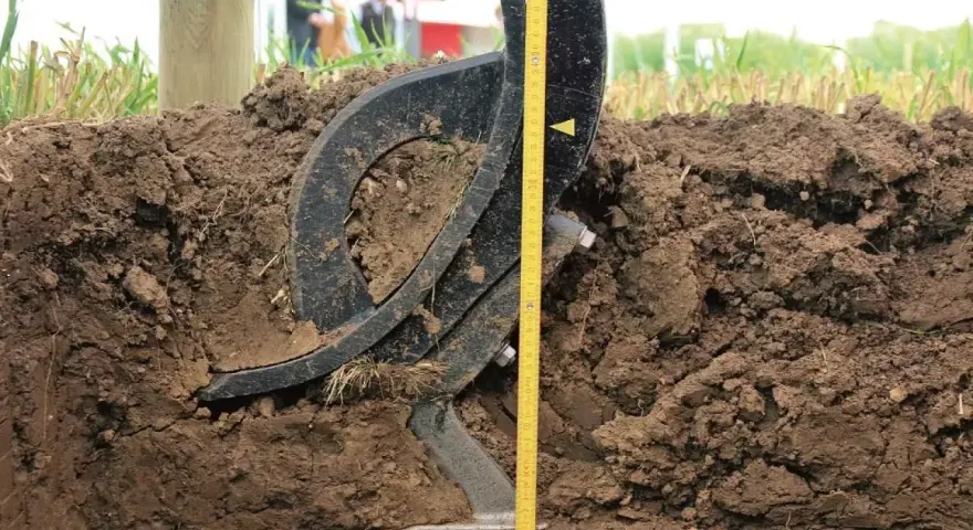 Czarne, metalowe elementy podeszwy płużnej zagłębiają się w ziemi. Obok miarka pokazuje głębokość orki.