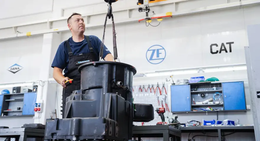 Mechanik steruje podnoszeniem dużych elementów skrzyni biegów w warsztacie. W tle logo ZF i CAT.