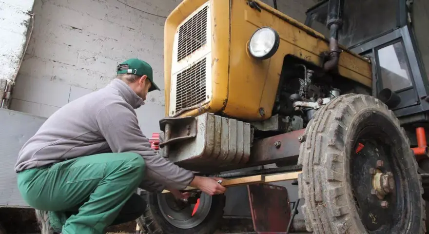 Mężczyzna w zielonych spodniach i czapce reguluje przednie koło żółtego ciągnika 4x4, pokazując, jak ustawić zbieżność.