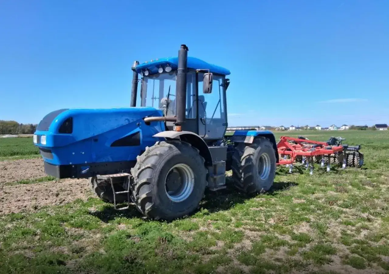 Niebieski traktor 100km z agregatem uprawowym na polu pod błękitnym niebem. Gotowy do pracy.