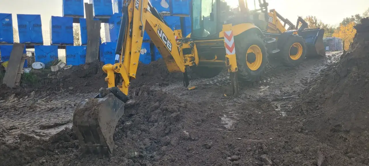 Ż&oacute;łta koparka JCB pracuje na błotnistym terenie. W tle widać palety z niebieskimi workami. Ile kosztuje godzina pracy koparki?