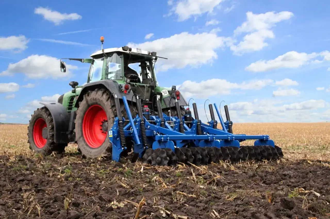 Zielony traktor z czerwonymi felgami wykonuje głęboką orkę na polu. Niebieskie narzędzie uprawowe rozbija ziemię, przygotowując ją do siewu.