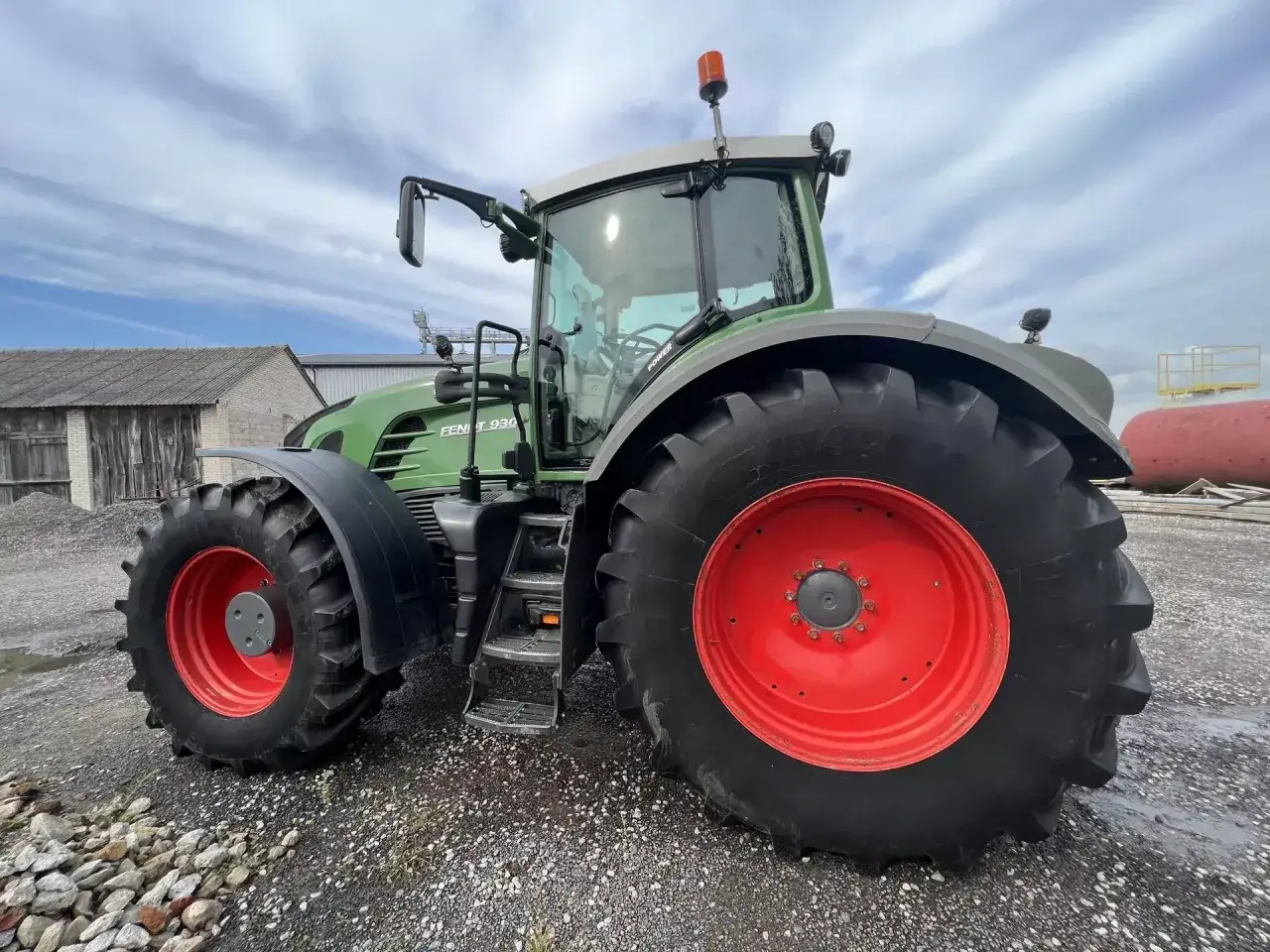 Zielony traktor Fendt 930 z czerwonymi felgami, stojący na żwirowej nawierzchni. Widoczne uszkodzenie maszyny - pęknięta opona.