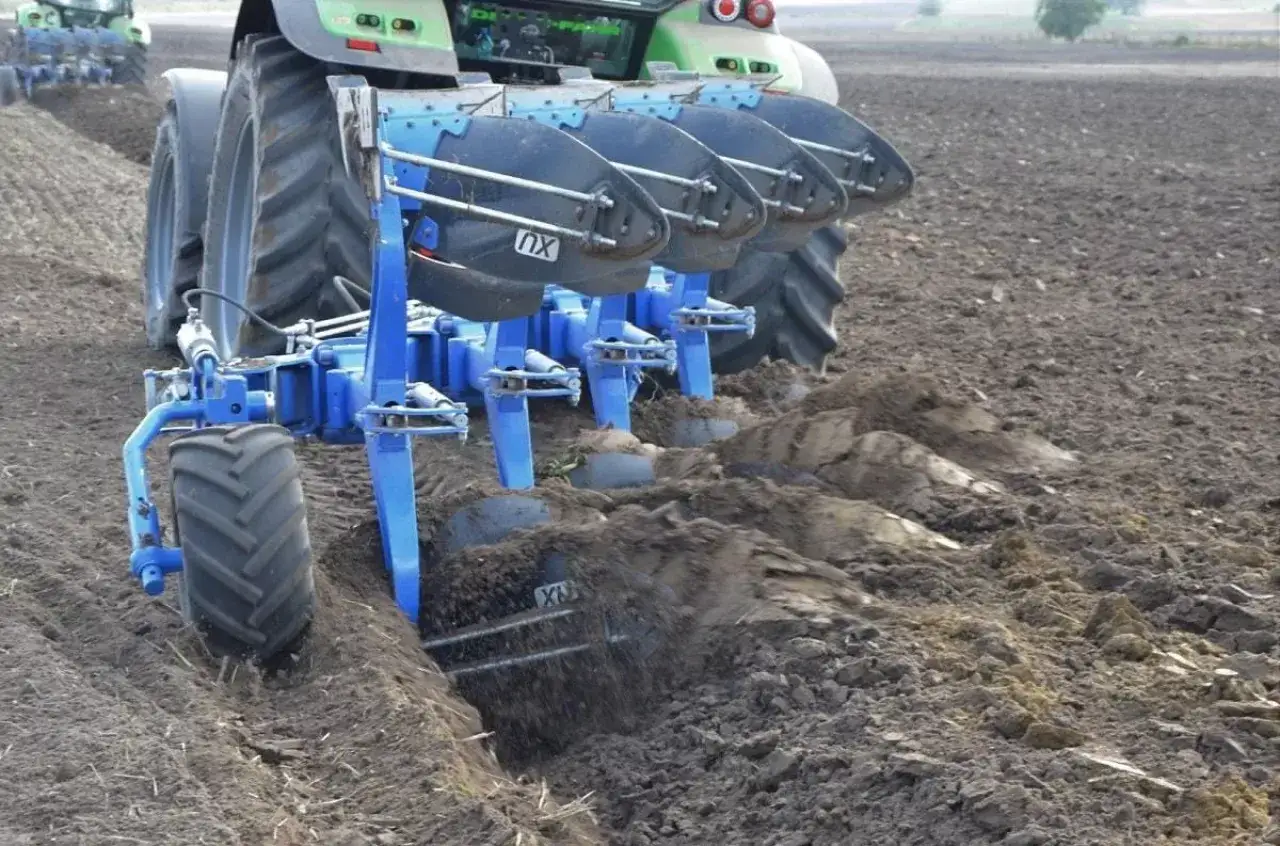 Traktor z pługiem obraca ziemię, pokazując, jak orać pole. Widać pracujące lemiesze i bryły ziemi.