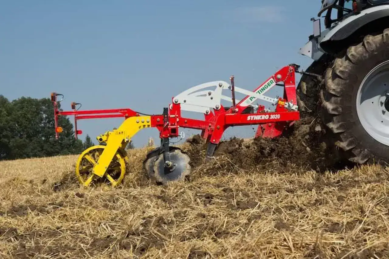 Czerwony kultywator Pöttinger Synkro 3020 pracuje na polu, spulchniając ziemię. To maszyna rolnicza do uprawy gleby.
