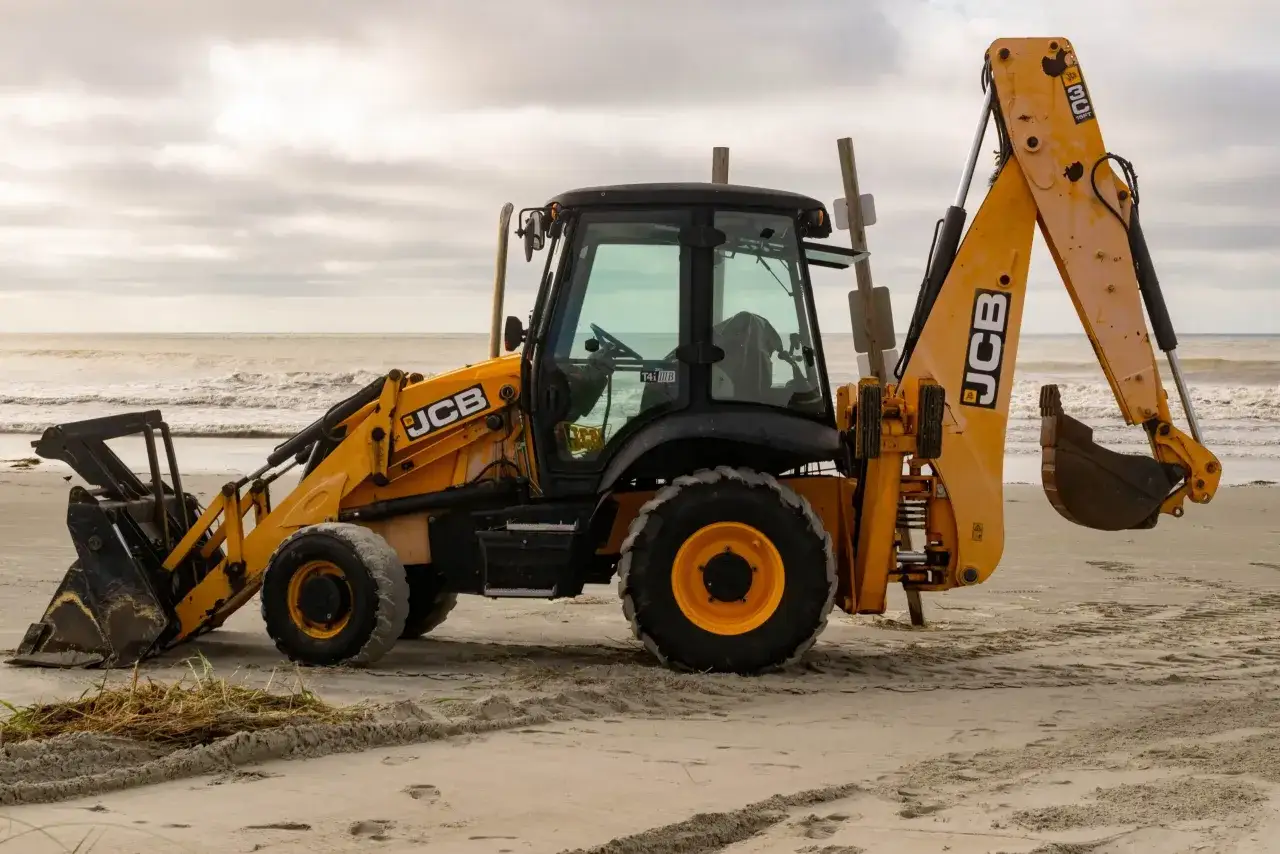 Ż&oacute;łty koparko-ładowarka JCB stoi na piaszczystej plaży, z widokiem na wzburzone morze i zachmurzone niebo.