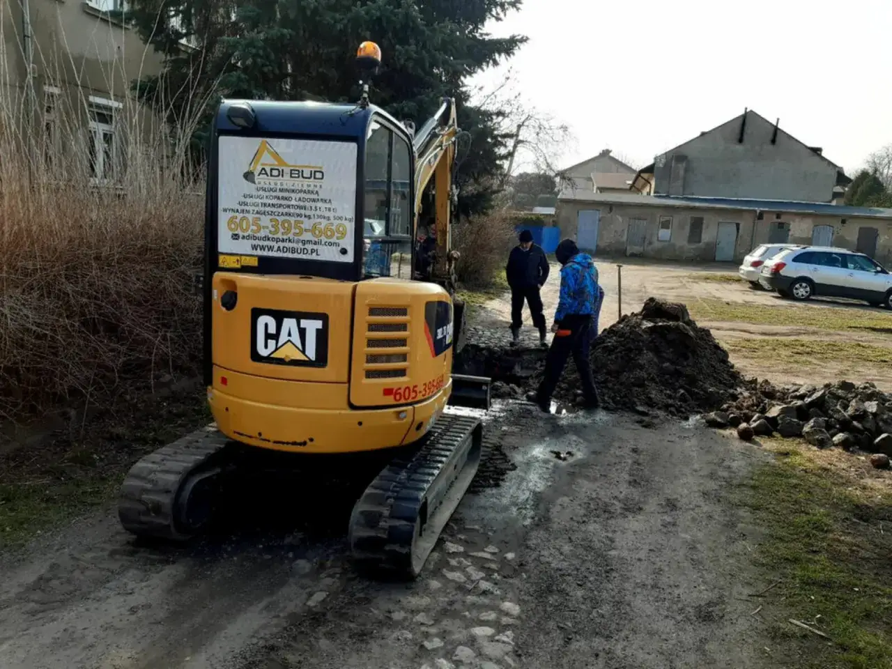 Minikoparka CAT pracuje przy wykopie. Na drzwiach maszyny widoczny numer telefonu i adres strony, gdzie można sprawdzić, ile kosztuje godzina pracy minikoparki.