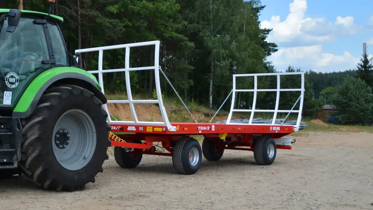 Zielony traktor ciągnie czerwoną przyczepę z białymi burtami. Przyczepa jest pusta, więc nie wiadomo, ile waży balot słomy, kt&oacute;ry m&oacute;głby ją wypełnić.