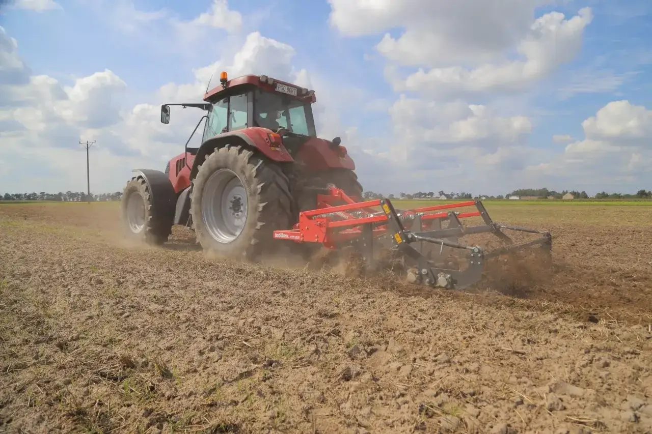 Czerwony traktor z podłączoną broną zębatą orze pole. Podeszwa płużna spulchnia ziemię, tworząc kurz.