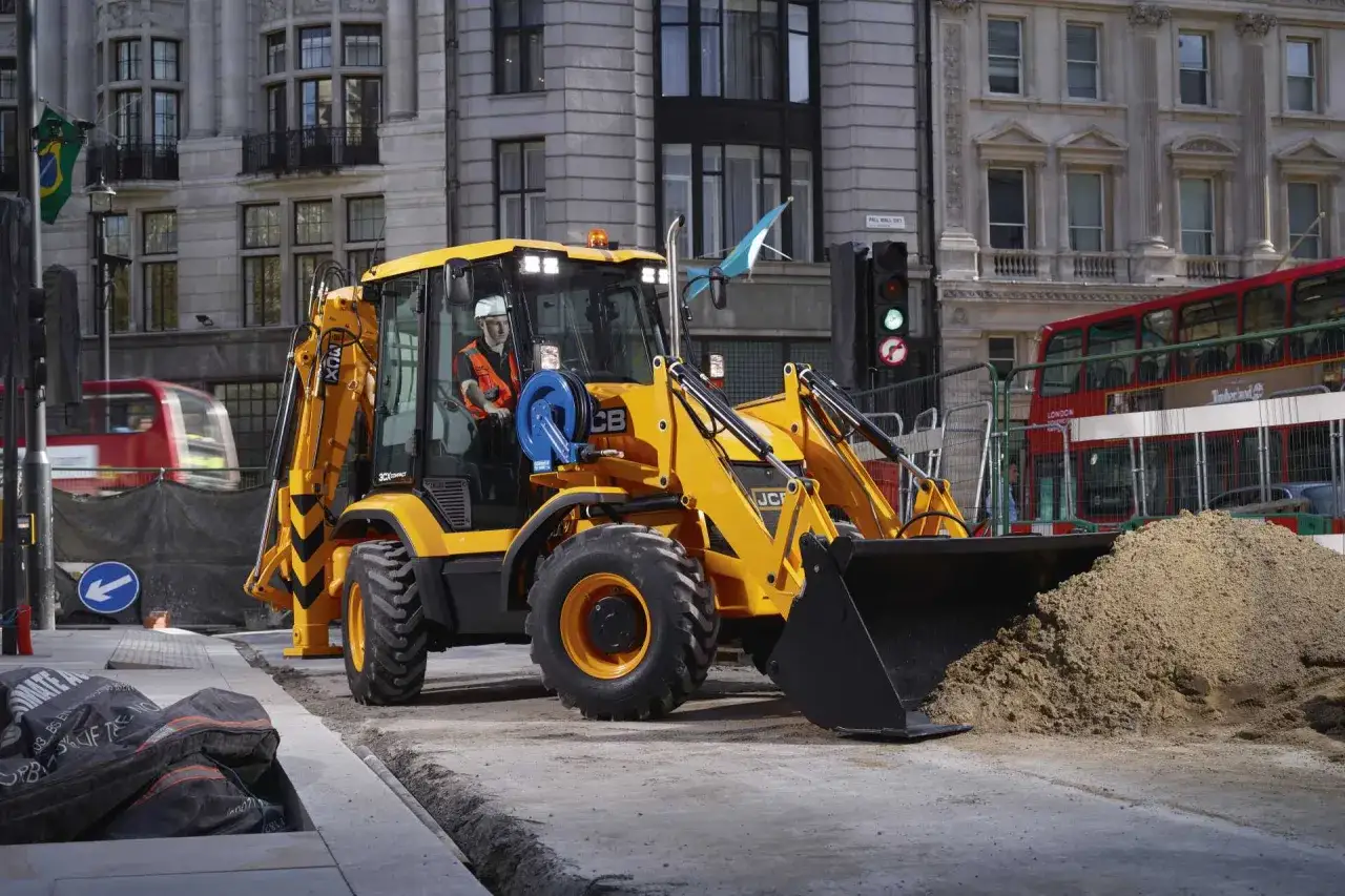 Żółta koparko-ładowarka JCB 3CX Compact pracuje na ulicy, ładując piasek. W tle widać zabytkową architekturę i czerwony autobus.