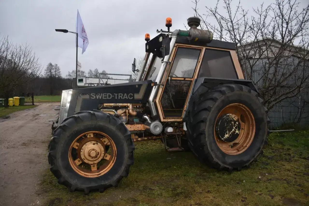 Ciemny traktor SWED-TRAC z pomarańczowymi światłami ostrzegawczymi stoi na trawie.