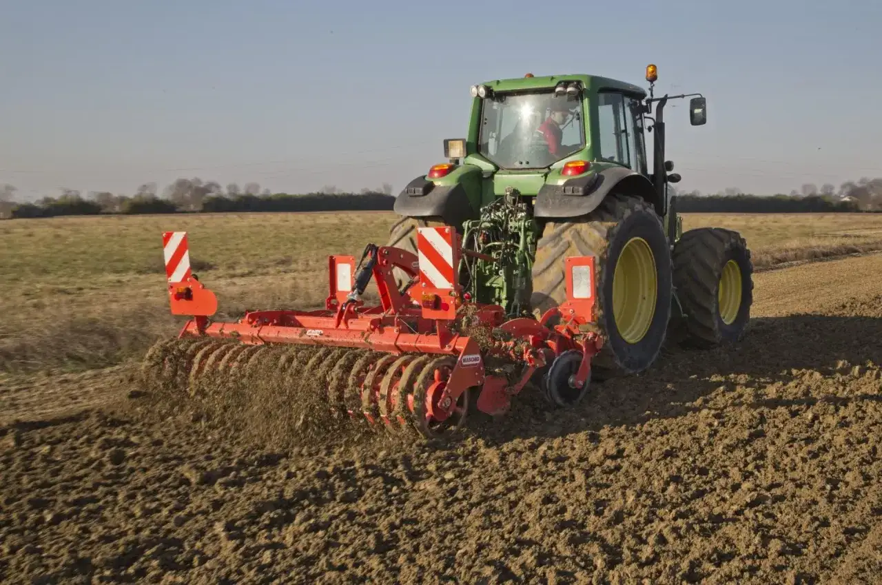 Zielony traktor z bronami wykonuje bronowanie po orce, spulchniając ziemię na polu.