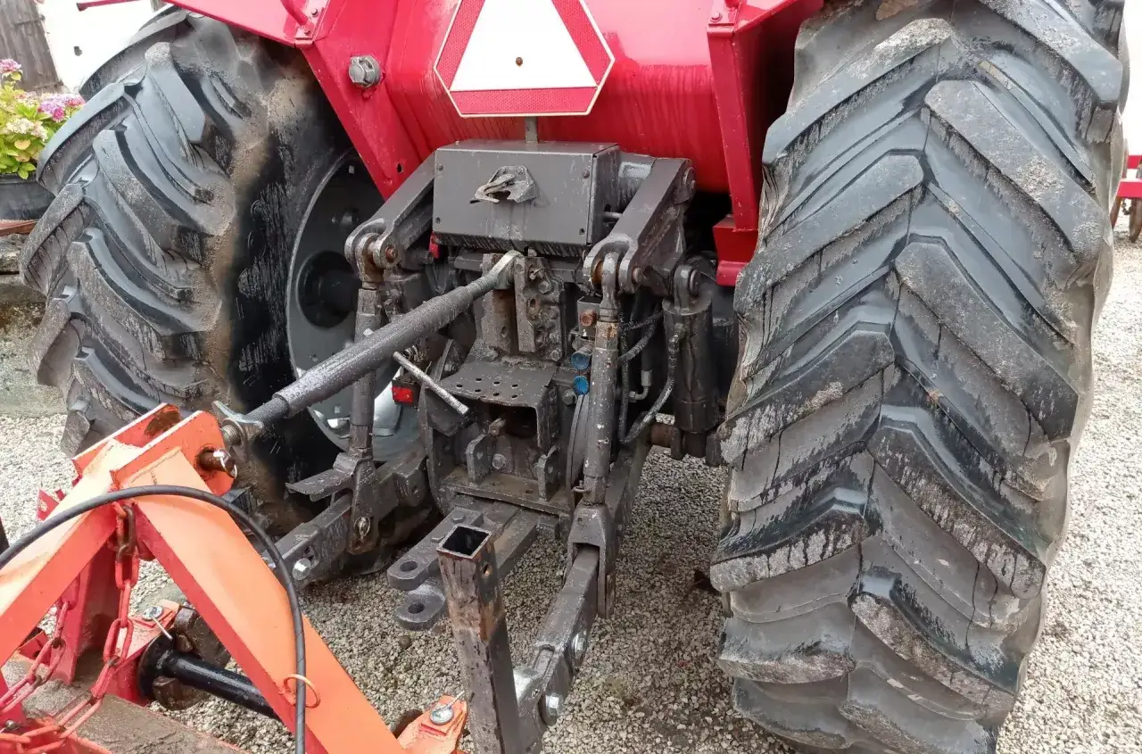 Czerwony traktor z dużymi, brudnymi oponami. Widać jego tylny podnośnik i zaczep. Może zaraz ruszy, ale obawiam się buksowania k&oacute;ł na żwirze.
