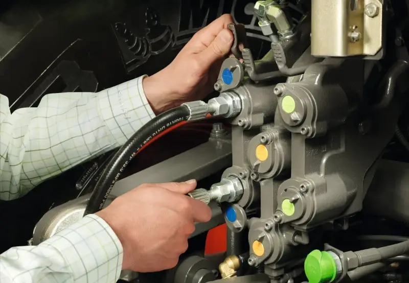 Ręce mechanika pracują przy skomplikowanym układzie hydraulicznym. Widać węże i zawory z kolorowymi oznaczeniami.