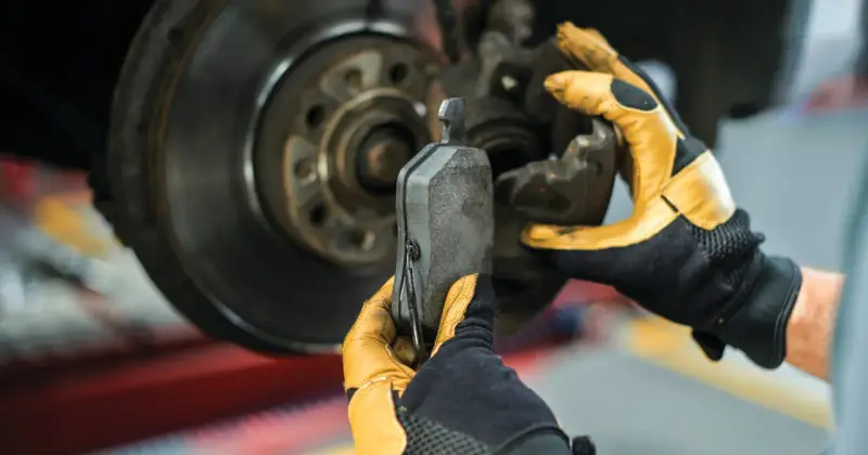 Mechanik w żółtych rękawicach wymienia klocki hamulcowe. Widoczny jest tarczowy układ hamulcowy.
